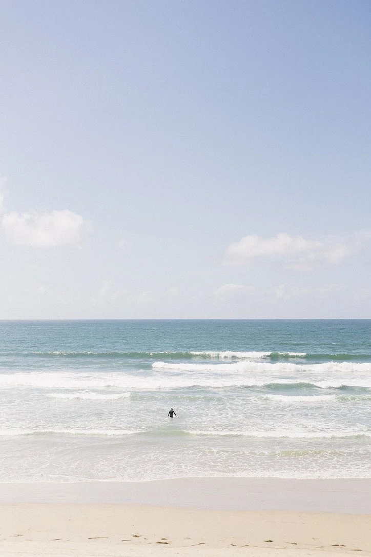 A person surfing in the ocean near the beach with blue sky and some clouds above.