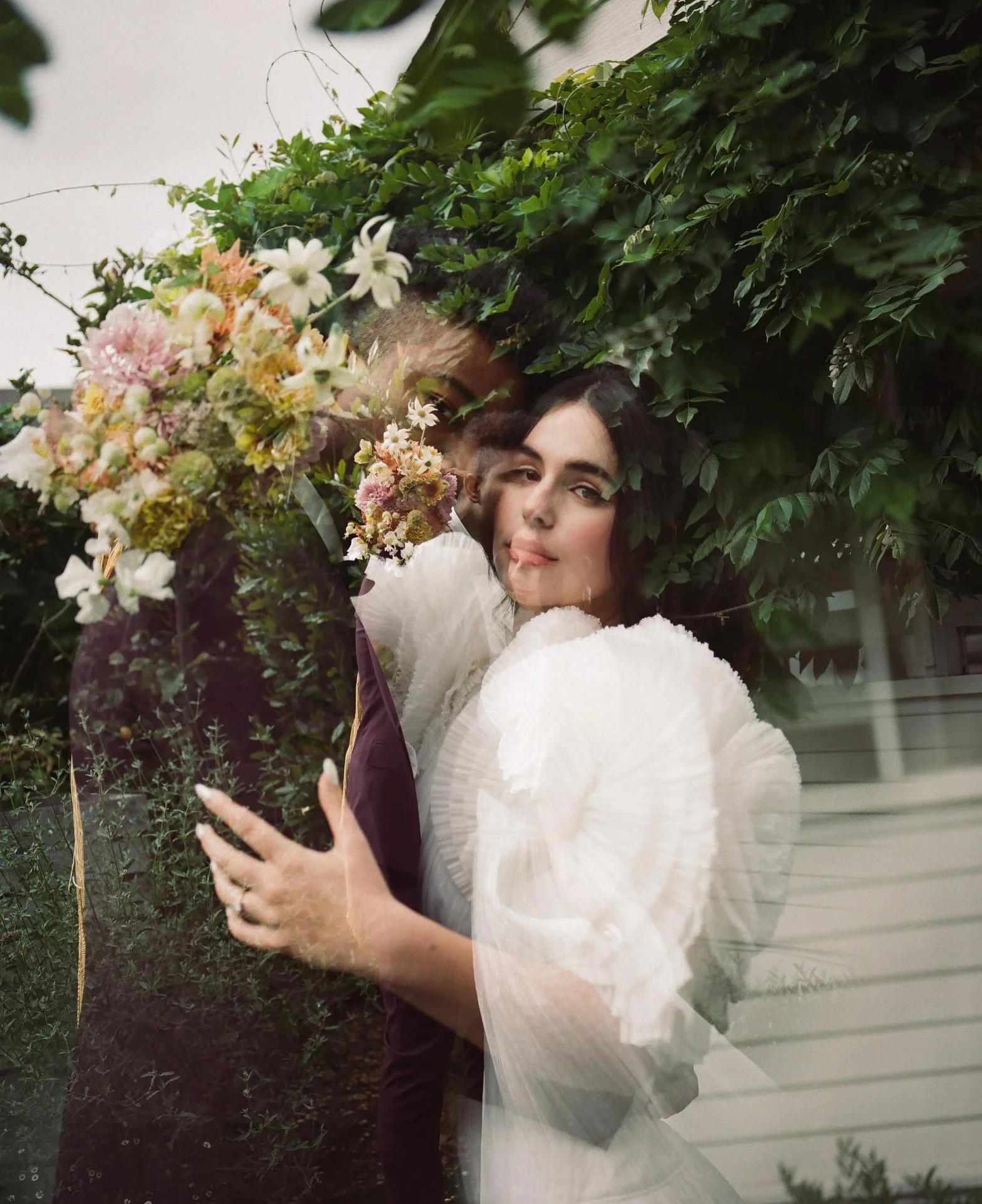 A woman and a man are looking through a glass window, with the woman holding a bouquet of flowers. The scene shows greenery outside the window, with the woman wearing a white dress and the man in a dark suit.