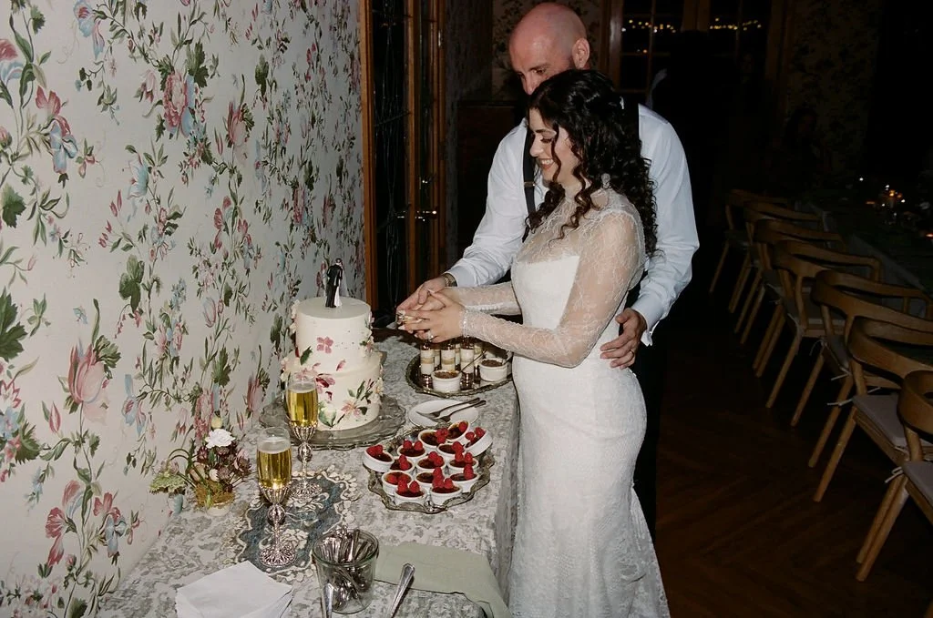 Bride and groom cutting wedding cake at reception with floral wallpaper and dessert table.