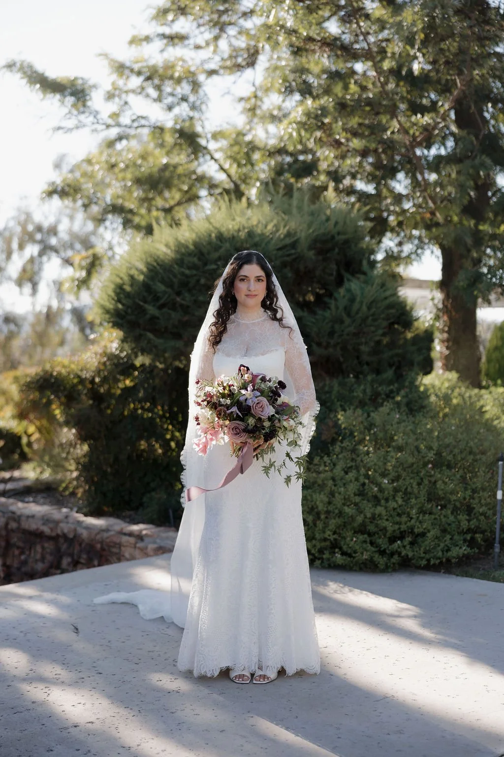 A bride in a white lace wedding dress standing outdoors with a bouquet of flowers, greenery, and trees in the background.
