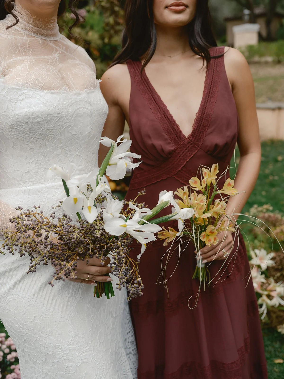 Two women in wedding and bridesmaid dresses holding floral bouquets outdoors.