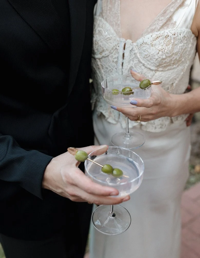 The image shows a close-up of two people holding elegant glasses with clear liquid and green olives on skewers, at a celebration or event.