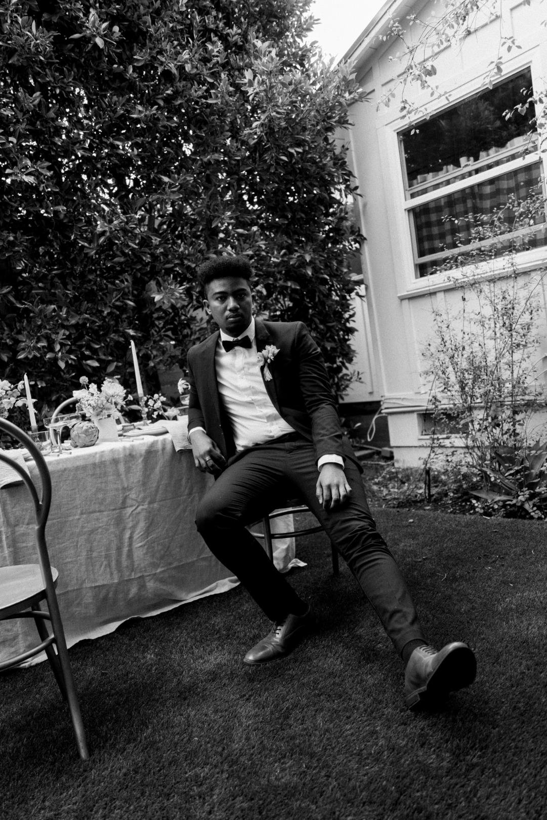 A young man in formal attire, wearing a tuxedo and bow tie, sitting on a chair outdoors. There is a garden with shrubs and a house in the background, and a table set for a meal nearby.