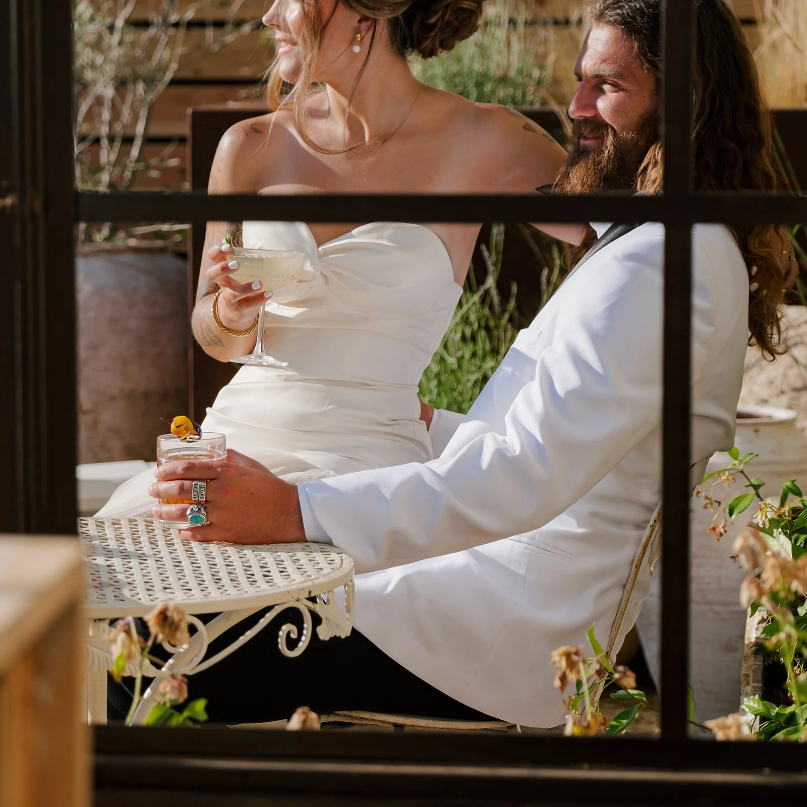 A woman in a white strapless dress and a man in a white suit jacket sitting outdoors, sharing drinks and smiling, seen through a window with metal bars.
