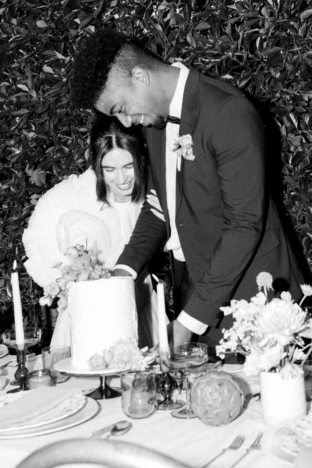 A couple at their wedding cutting a cake, with the groom in a tuxedo and the bride in a white dress, surrounded by floral decorations and candles.