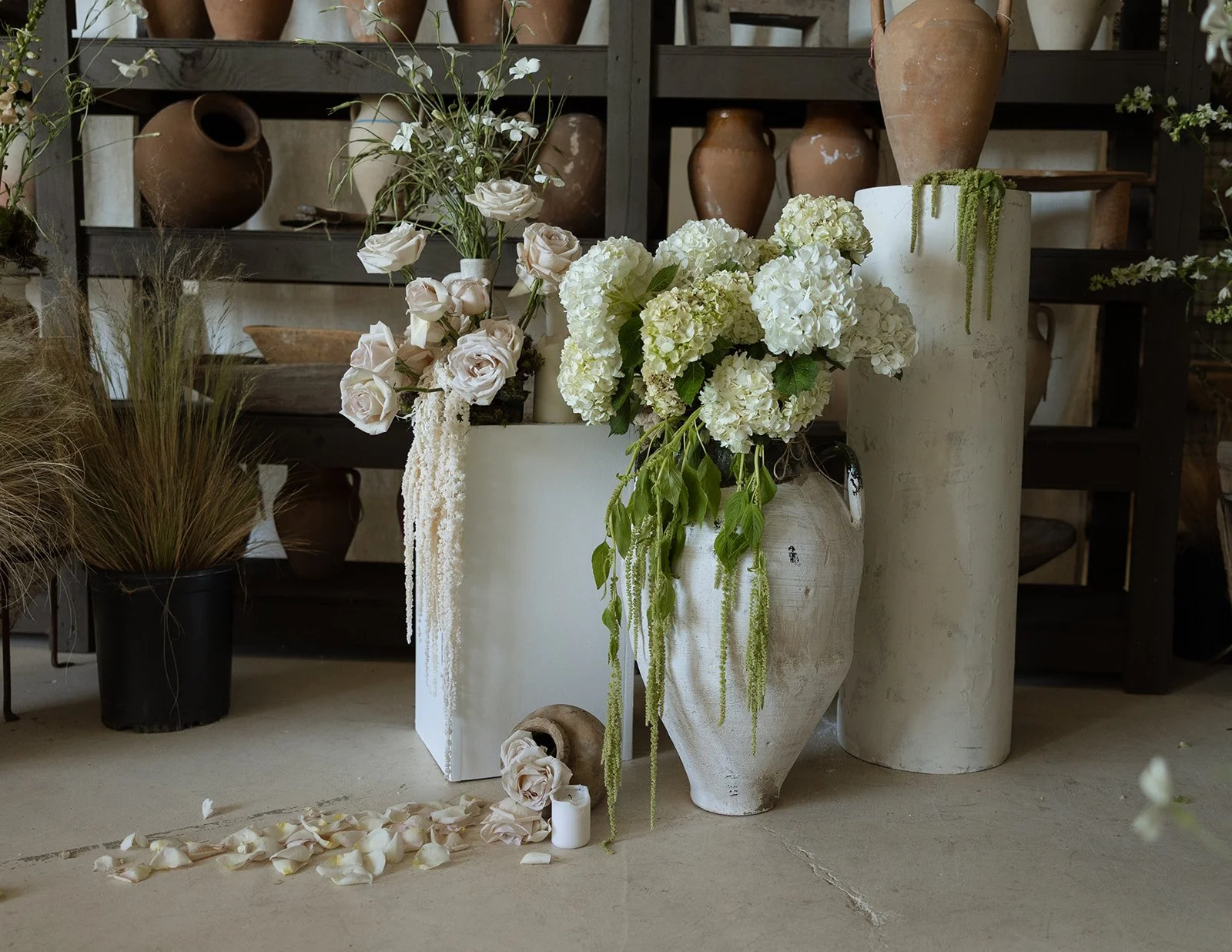 Vases with white and pale pink flowers, flower petals scattered on the floor, and pottery in a rustic setting.