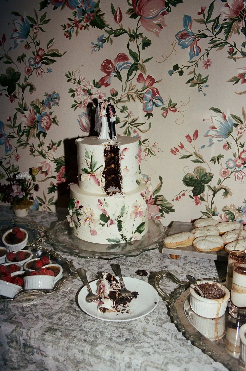 Wedding cake with floral decorations, partially cut to reveal chocolate layers, surrounded by desserts like berries, cookies, and puddings on a decorated table, with floral wallpaper background.