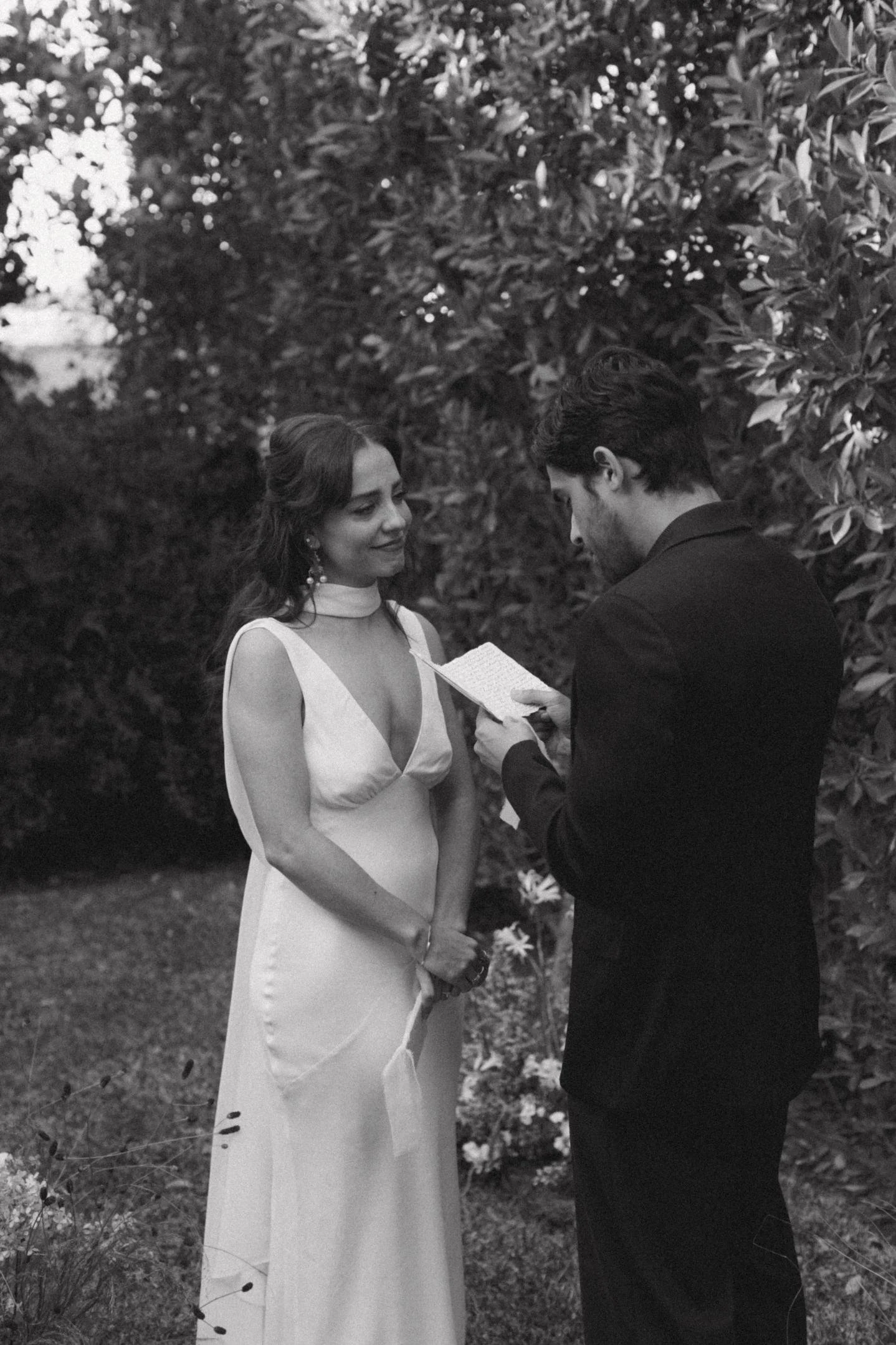 A man and woman exchange vows outdoors, with the man reading from a small notebook. The woman wears a white dress with a high neckline and veil, standing in front of dense foliage.