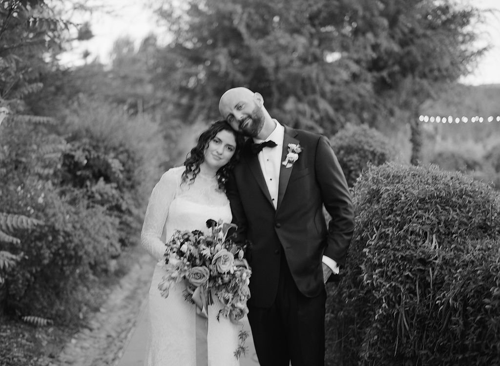 Black and white photo of a bride and groom standing outdoors near bushes and trees, with the groom leaning his head on the bride's head and they smiling gently.