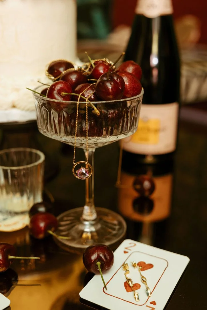 A glass bowl filled with cherries and jewelry, with a playing card and a bottle in the background, on a reflective surface.