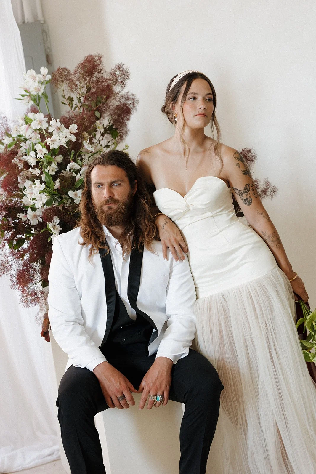 A man with long hair and a beard, wearing a white suit with a black shirt and tie, seated beside a woman in a white strapless dress, against a background of large floral arrangements.