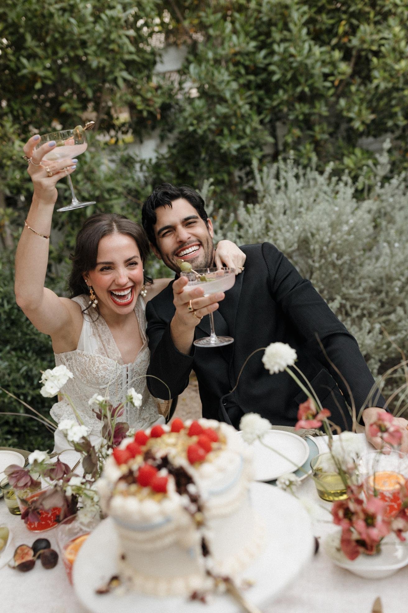 A happy couple celebrating at a wedding or party, raising glasses with drinks, sitting at a decorated table with a cake and flowers outdoors.