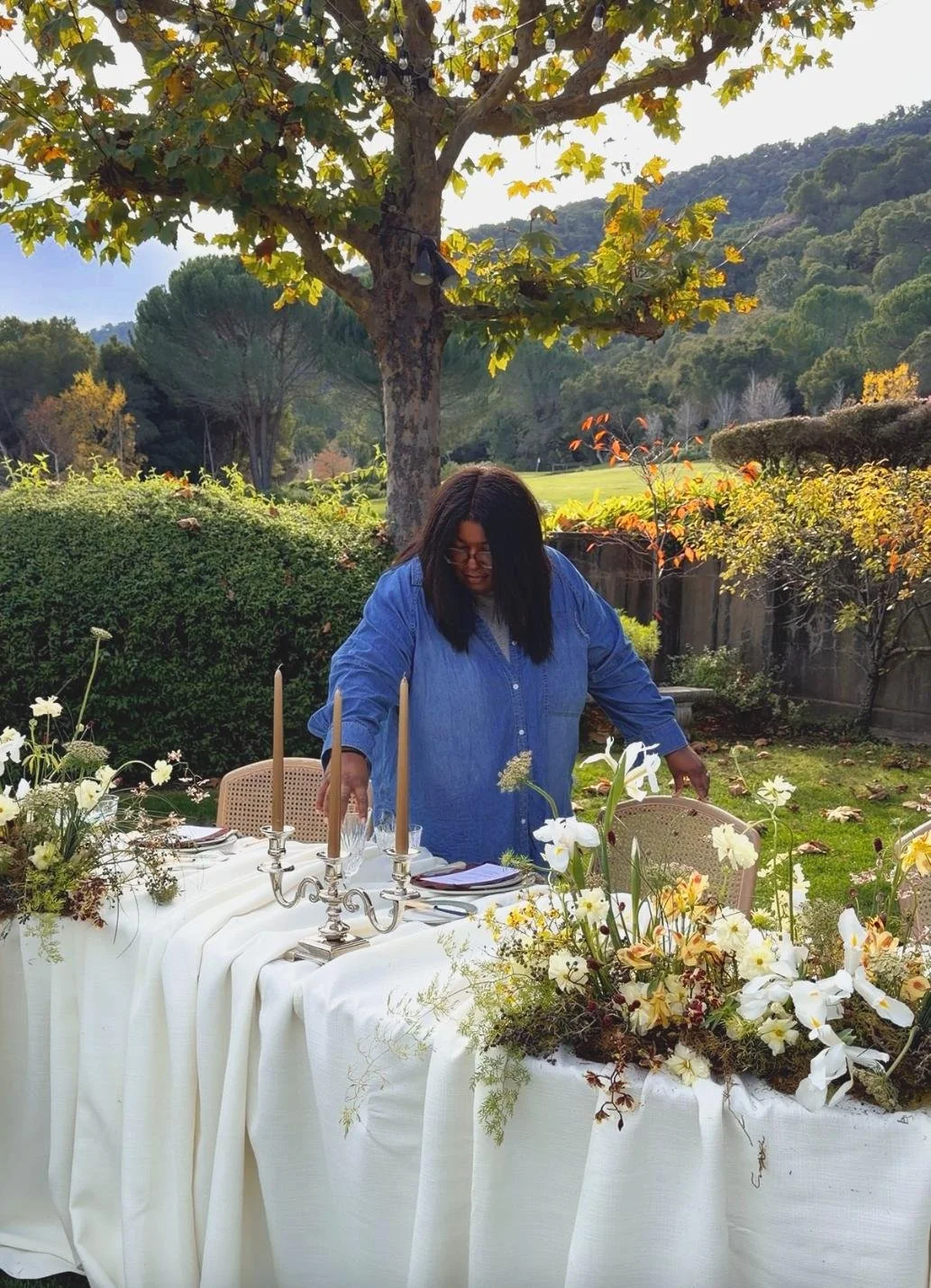 A person in a blue shirt arranging an outdoor dining table decorated with white flowers and candles under a tree in a garden.