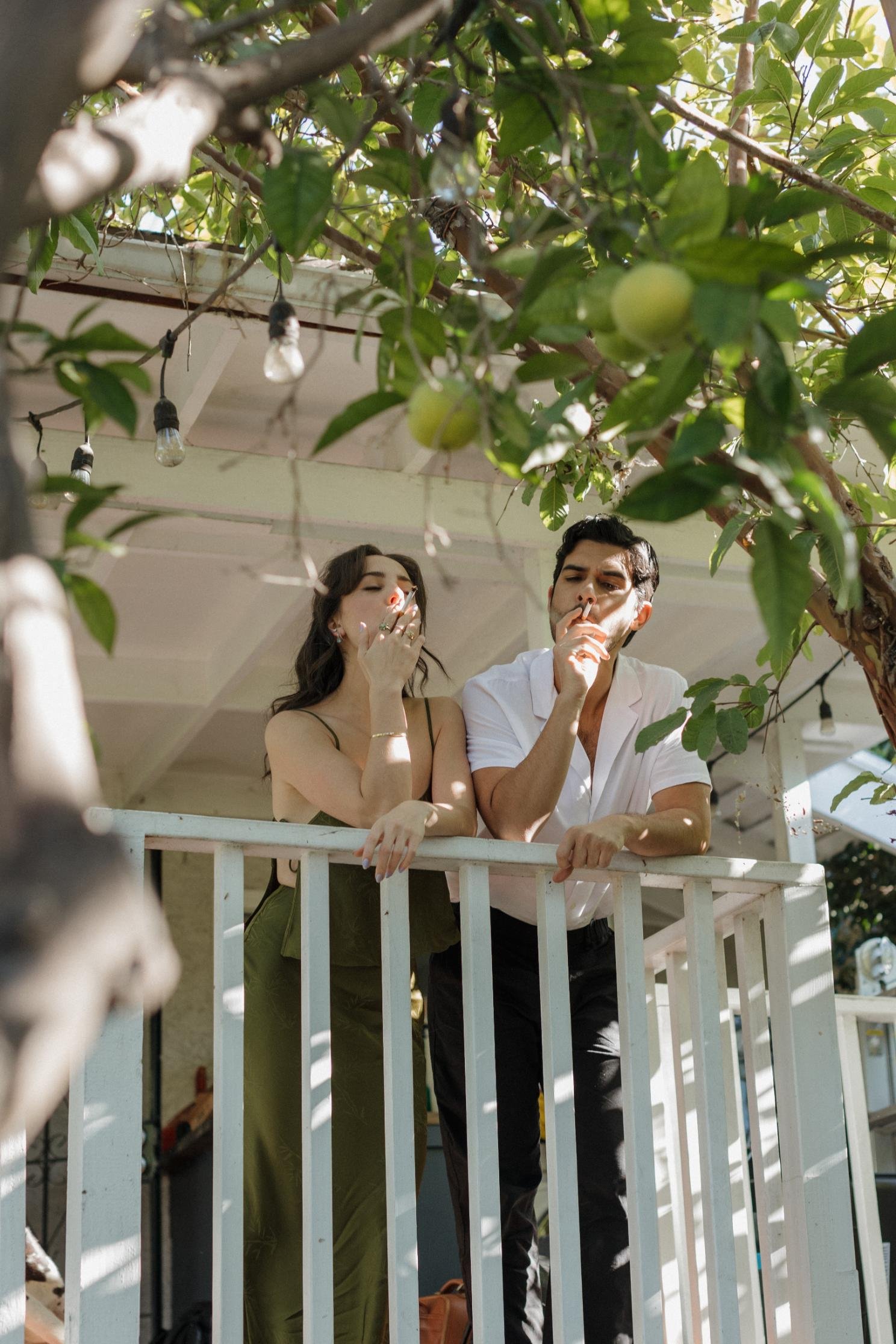 A man and woman smoke cigarettes on a balcony under a tree with green apples, with string lights overhead.