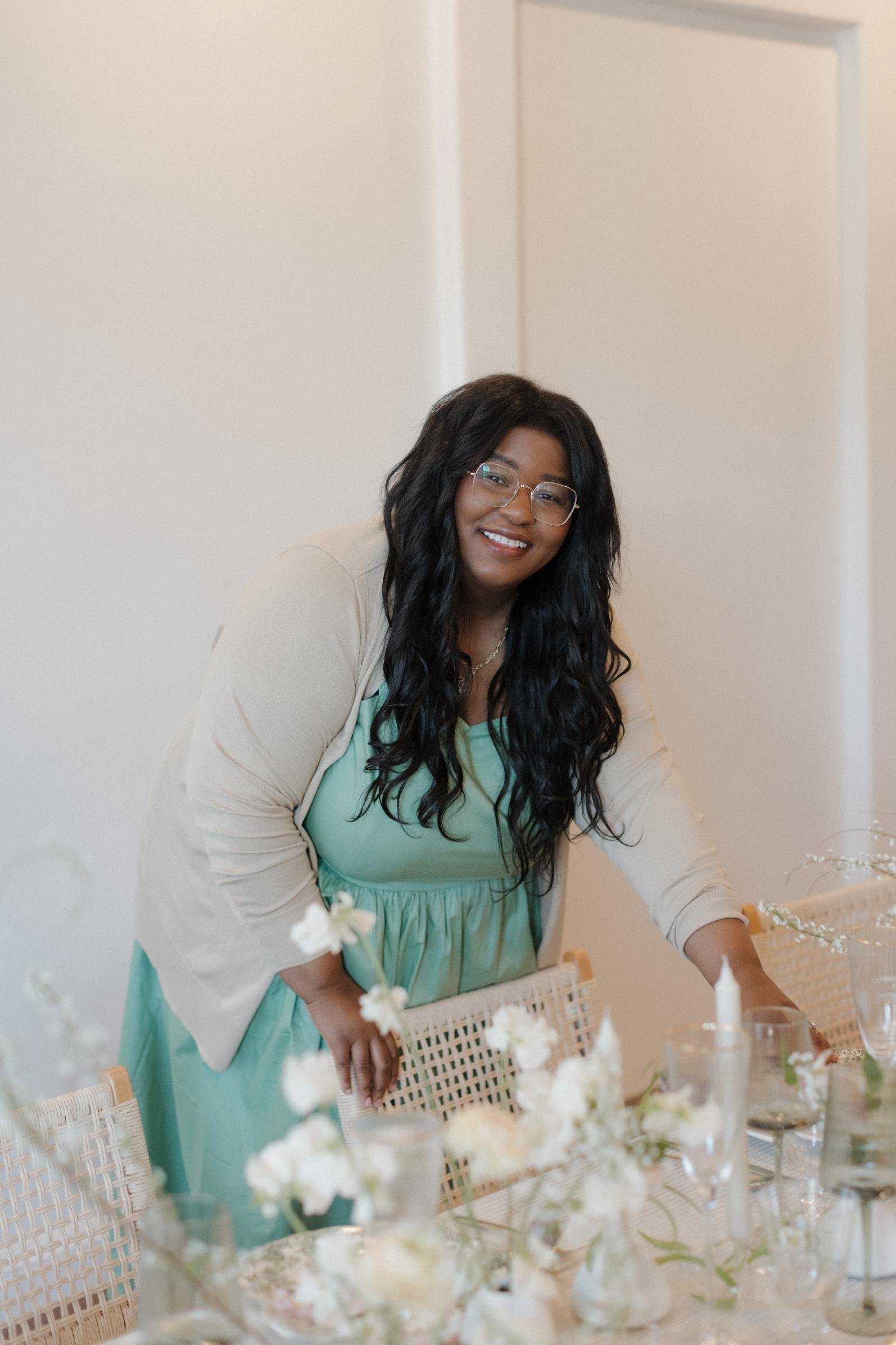 Averi Linch Wedding Planner A woman with long curly black hair and glasses smiling, leaning over a table with floral arrangements, in a well-lit room.