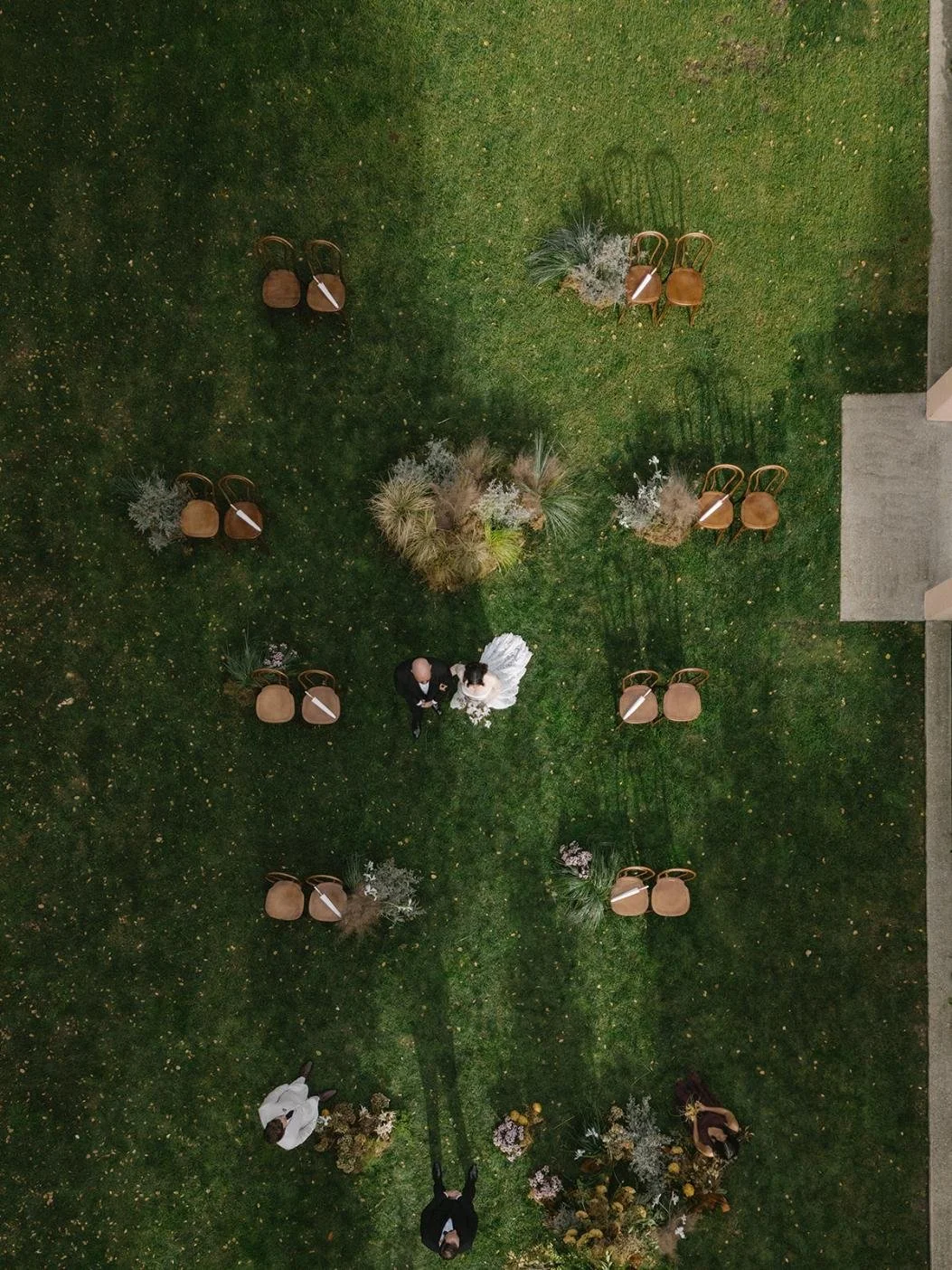 An aerial view of a wedding ceremony outdoors with chairs arranged on a grassy area, flowers, and a bride and groom standing in the center.