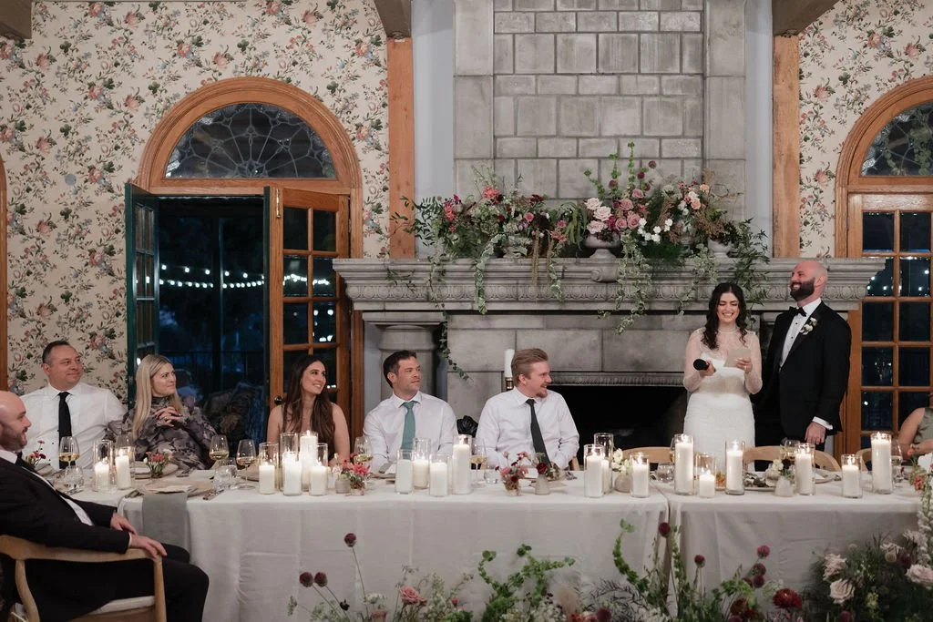 A wedding reception with a head table where the newlywed couple is standing and giving a speech, surrounded by seated guests in a decorated room with floral wallpaper, large windows, and a stone fireplace topped with flowers and greenery.