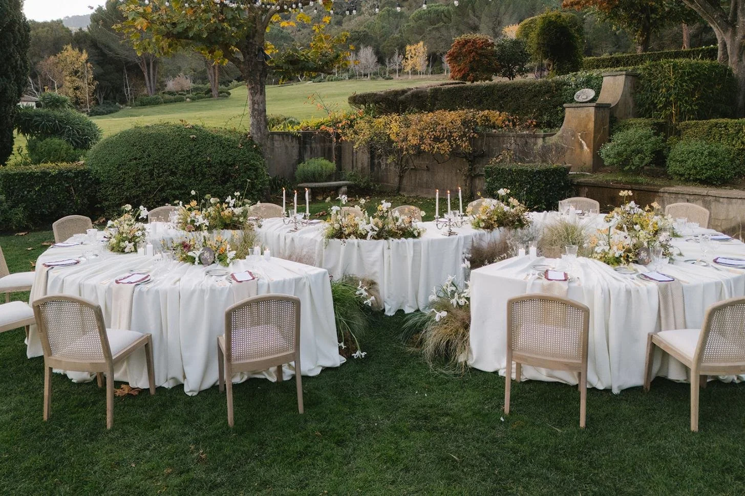 Outdoor wedding reception with round tables draped in white tablecloths, decorated with floral centerpieces, candles, and place settings, set in a lush garden with green grass, trees, and foliage in the background.