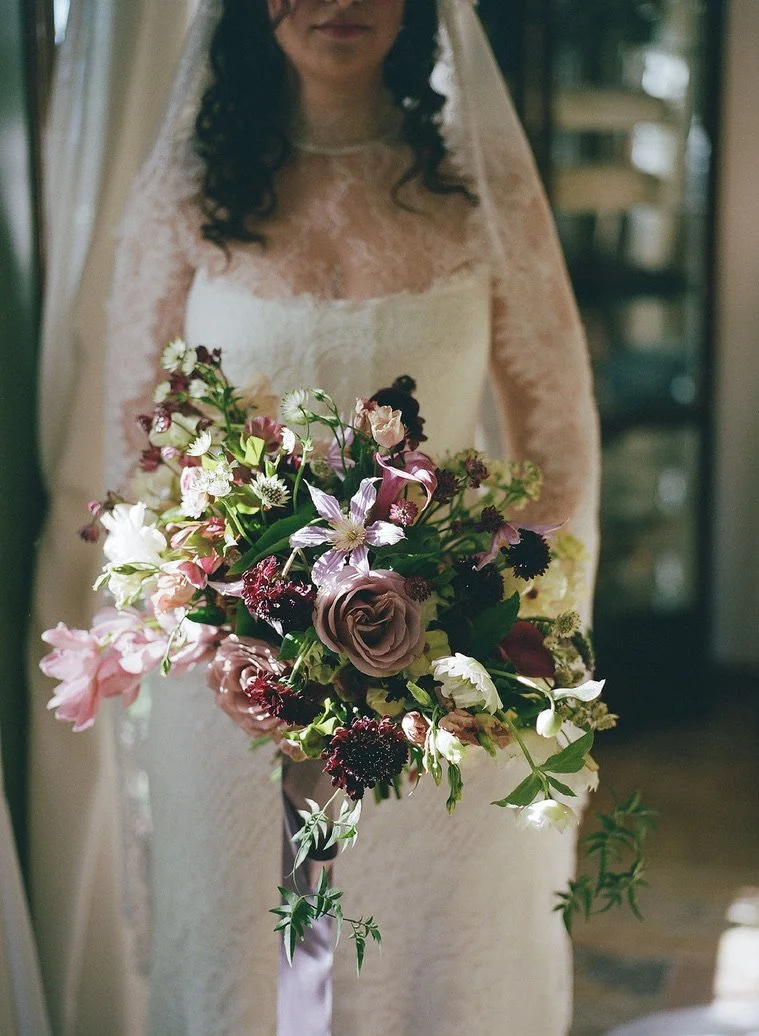 Woman in a lace wedding dress holding a bouquet of mixed pink, purple, and white flowers with green foliage.