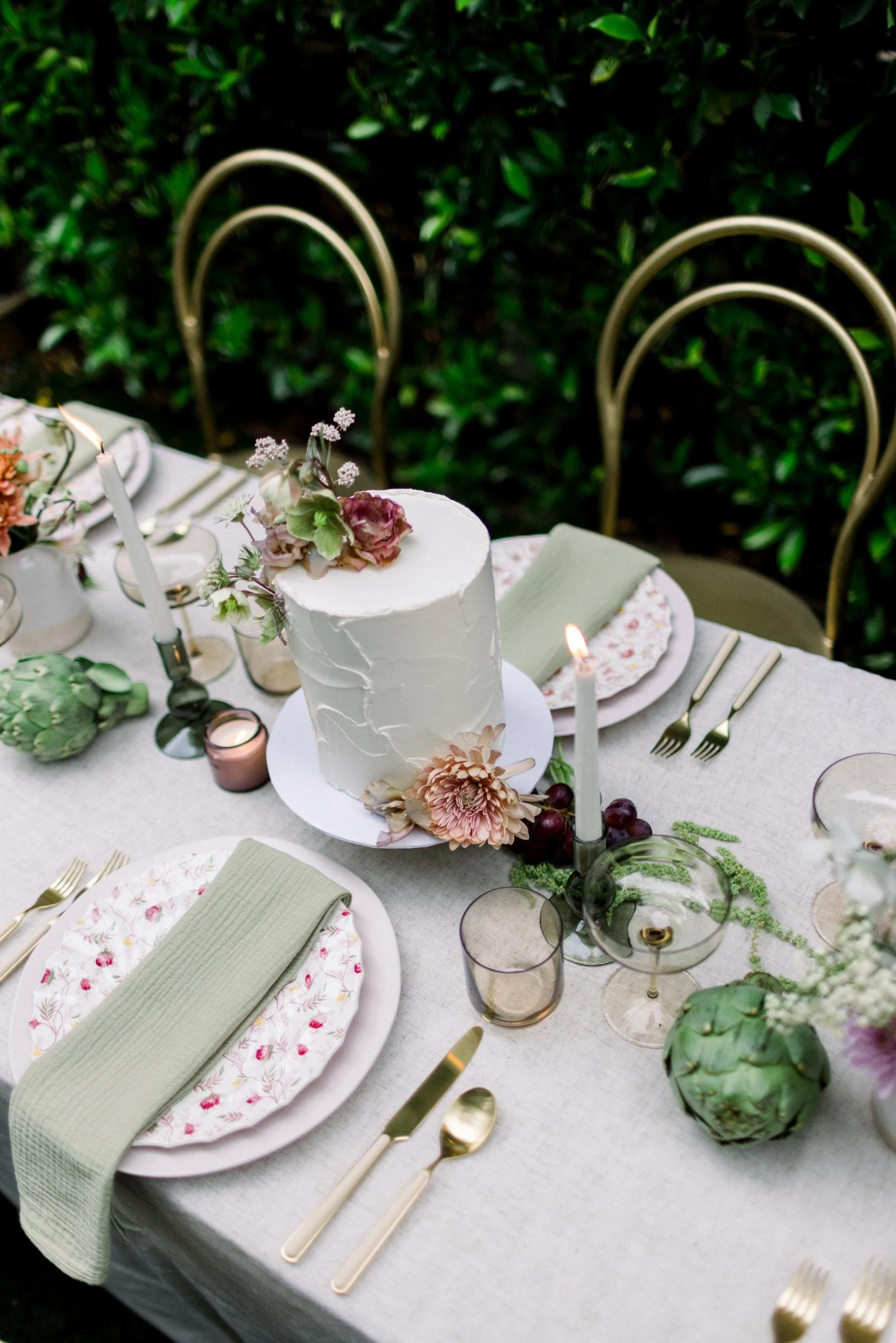Elegant outdoor table setting with a white frosted cake decorated with flowers, green foliage, candles, and gold flatware, surrounded by chairs and greenery in the background.