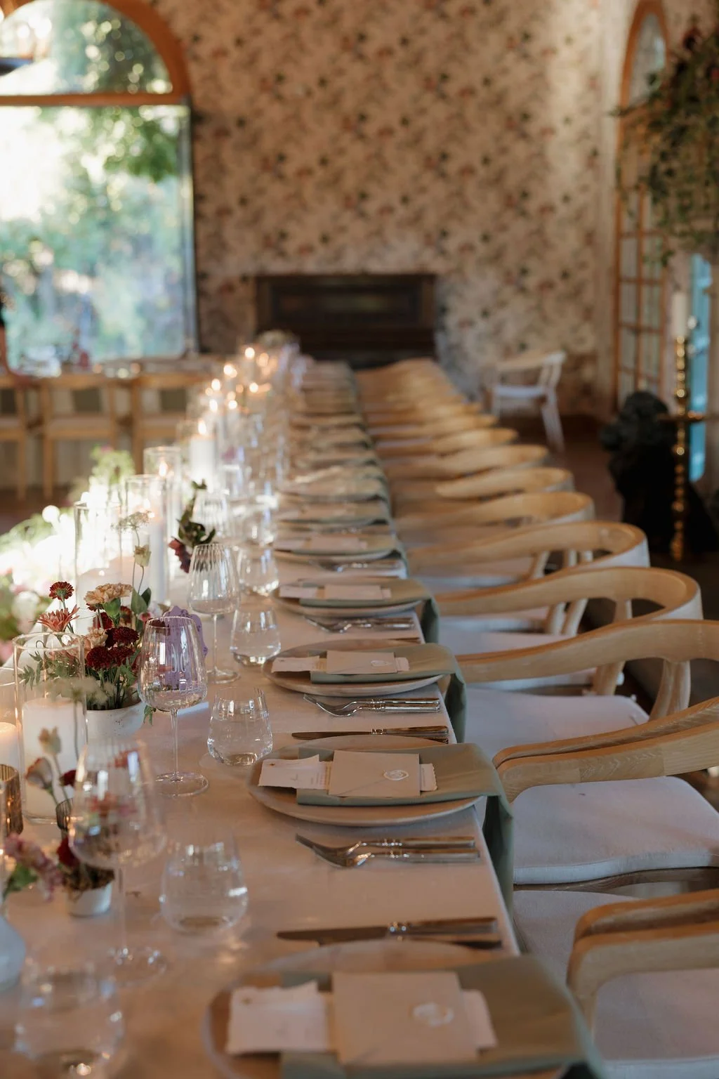 A long dining table set for a wedding or special event with floral centerpieces, candles, wine glasses, and place settings in a decorated room with large windows and floral wallpaper.