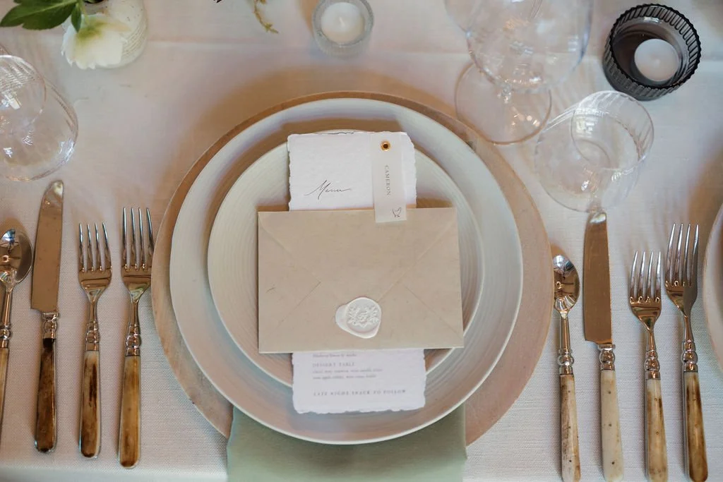 Table setting with a beige charger plate, white dinner plate, napkin, and an envelope with a wax seal, surrounded by multiple glasses and silverware, on a white tablecloth.