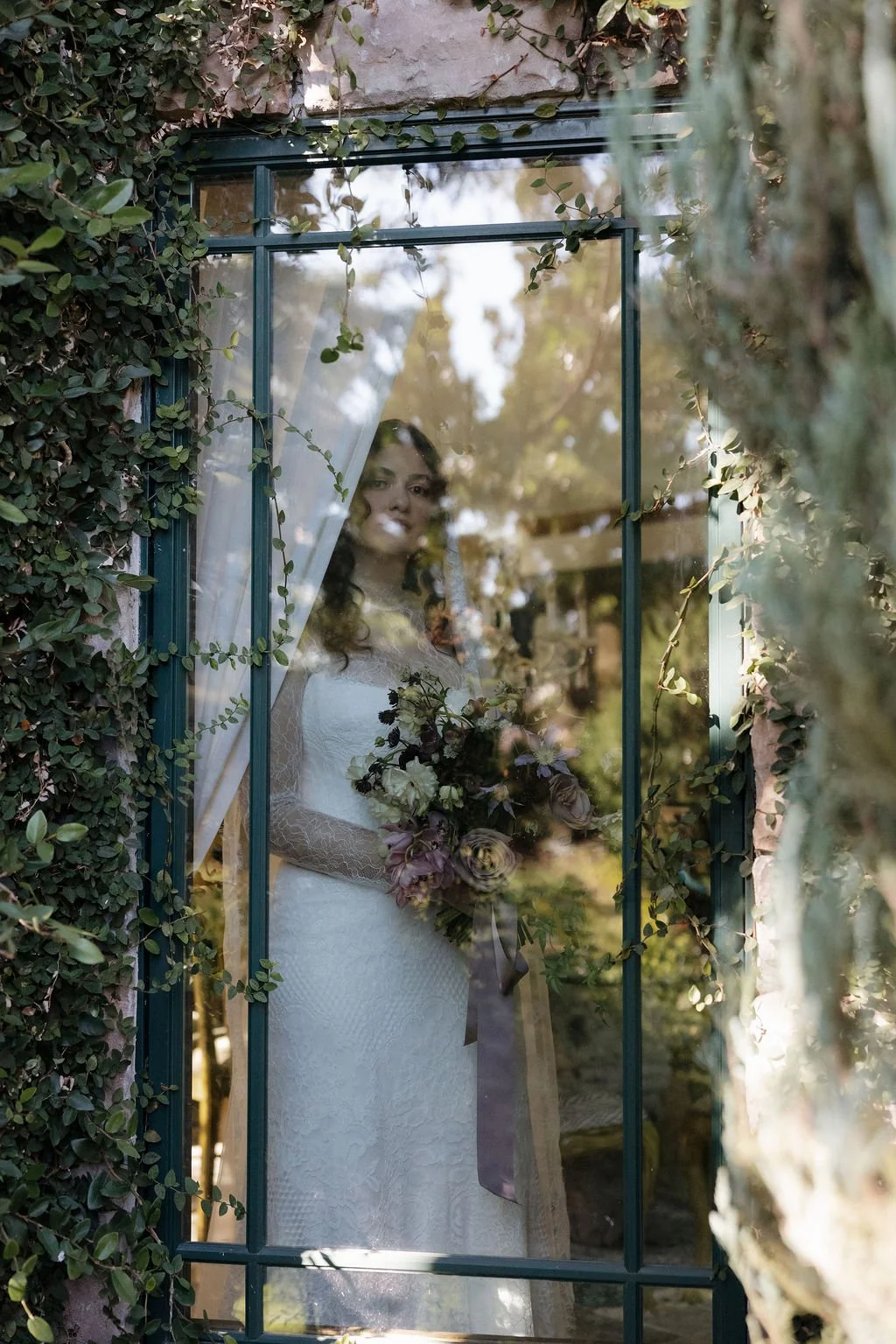 A woman in a white wedding dress holding a bouquet of flowers, seen through a glass window with reflections of trees and surrounding foliage, framed by ivy-covered walls.