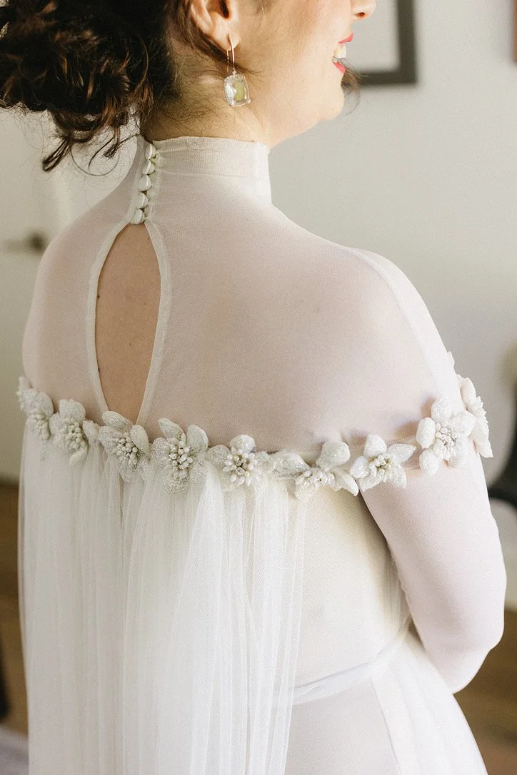 A woman wearing a white dress with floral embellishments and a keyhole back, seen from behind, with earrings and curly hair styled in an updo.