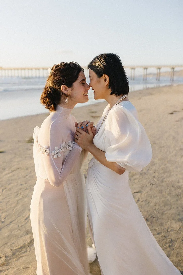 Two women in white dresses holding hands and touching foreheads on a beach with a pier in the background.