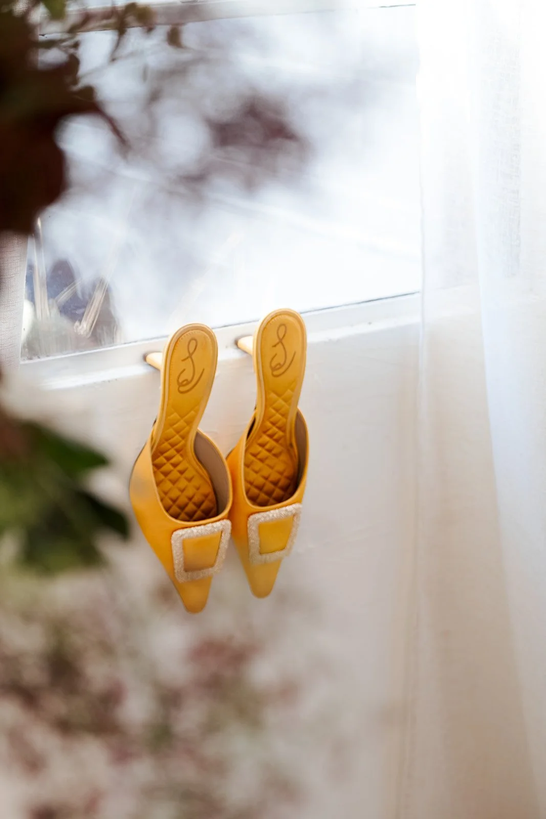 Pair of yellow high-heeled shoes with square rhinestone buckles hanging on a windowsill.