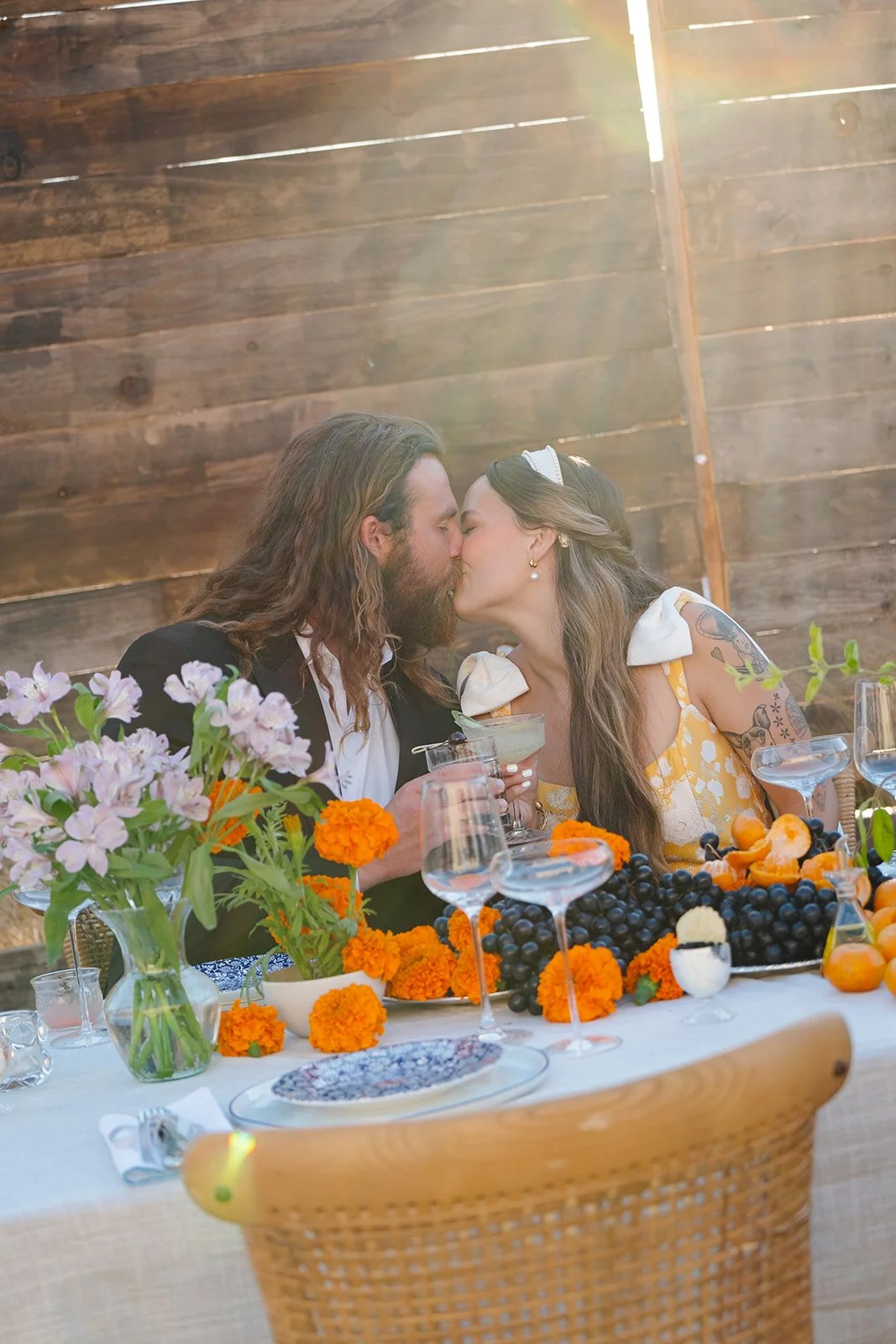 A couple sharing a kiss at a decorated table with flowers and fruit during a celebration.