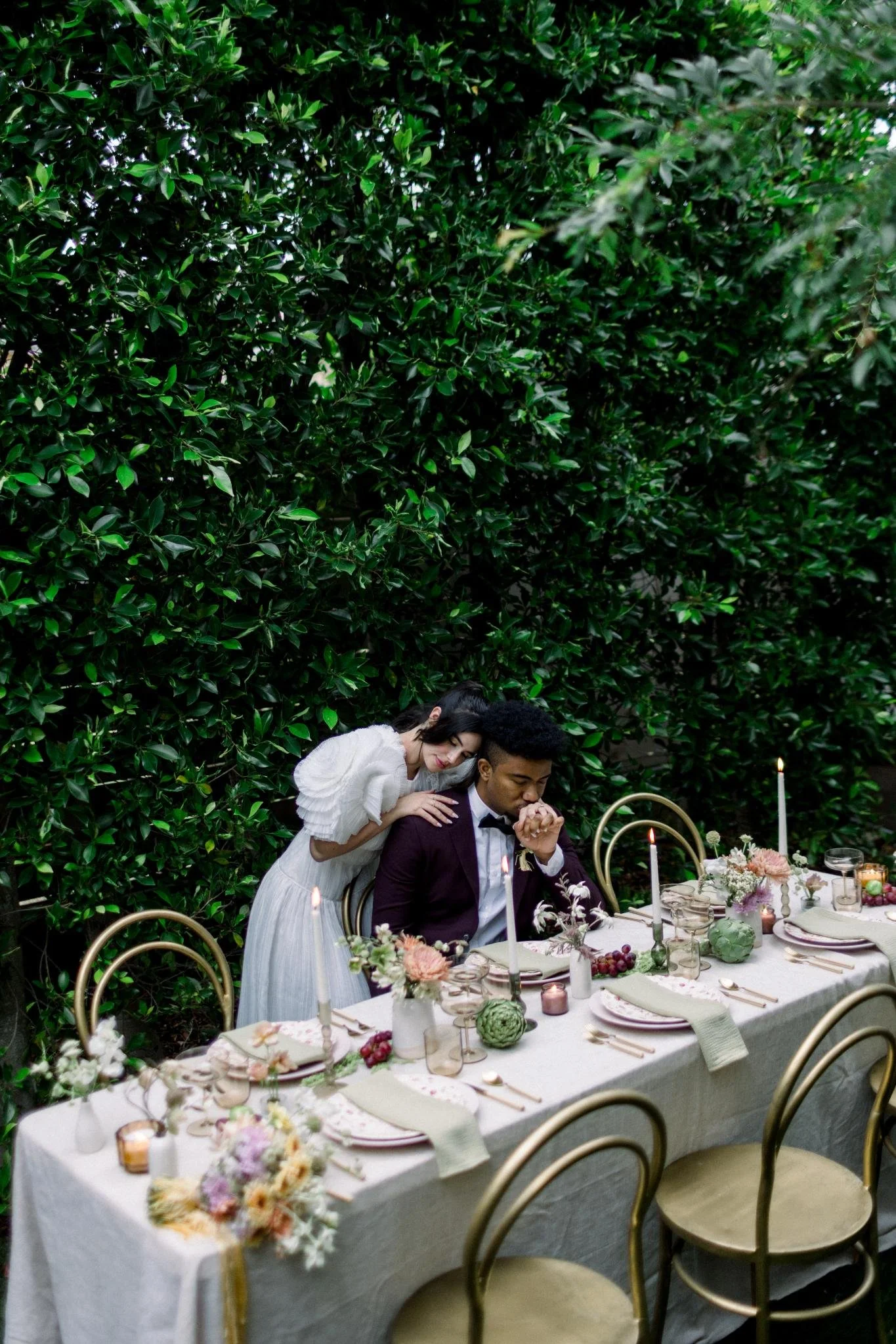 A couple dressed formally, with the woman in a white dress leaning on the man's shoulder at a decorated dining table outdoors.