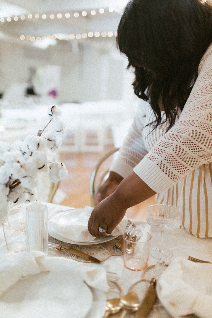 Averi Linch Wedding Planner A woman arranging table settings for a formal event or celebration, with cotton branch centerpiece, gold and pink accents, and elegant glassware.