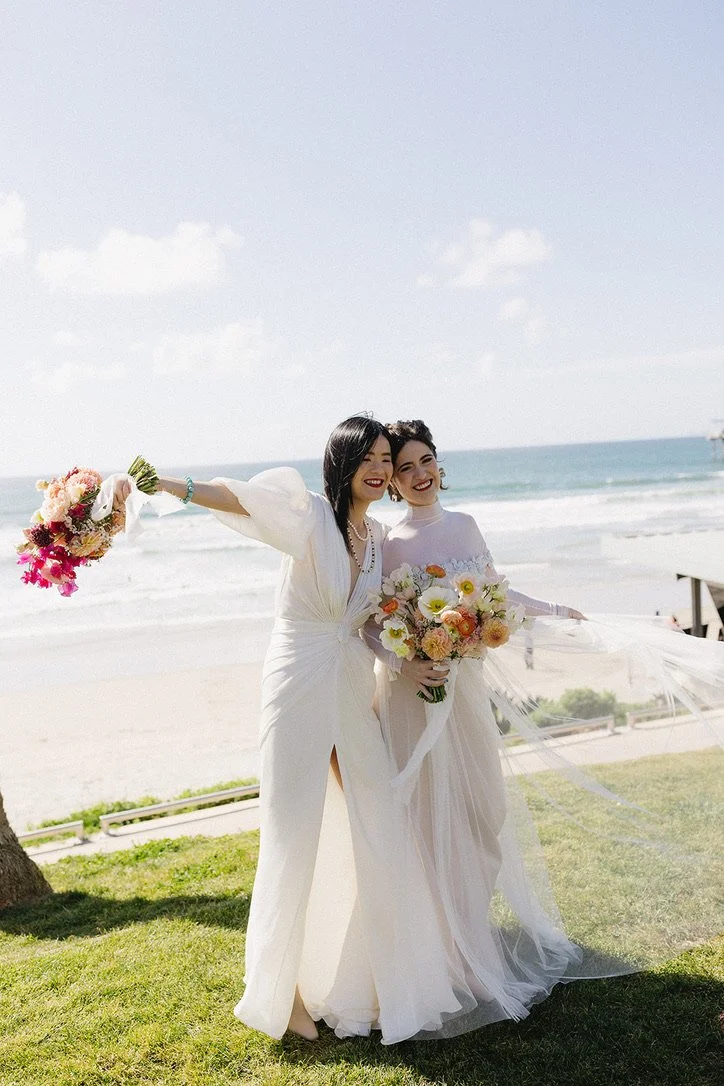 Two women in wedding dresses holding bouquets, standing on grass near the beach with ocean waves and a cloudy sky in the background.