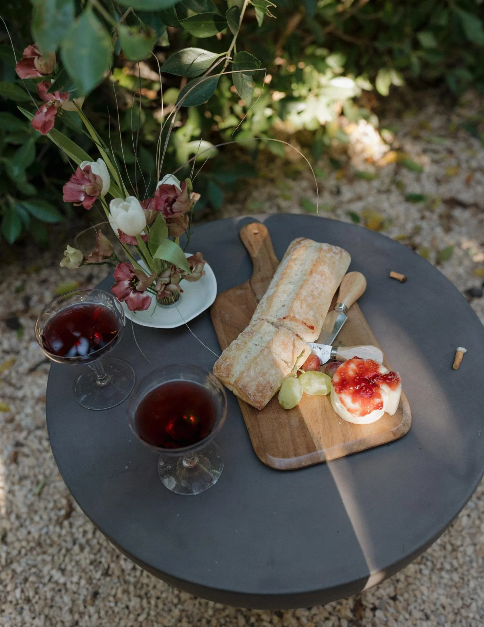 A black outdoor table with a cheese and bread platter, two glasses of red wine, and a pink and white flower arrangement.