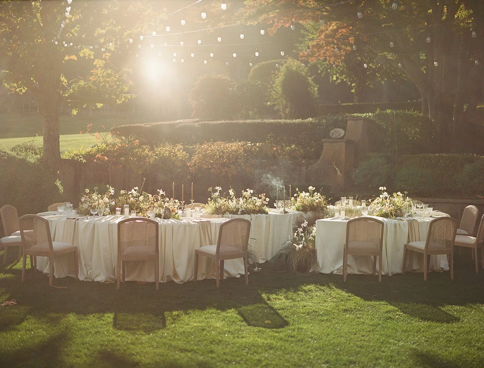 Elegant outdoor wedding reception setup with round tables draped in white tablecloths, decorated with floral centerpieces, set in a lush garden with sunlight filtering through trees.