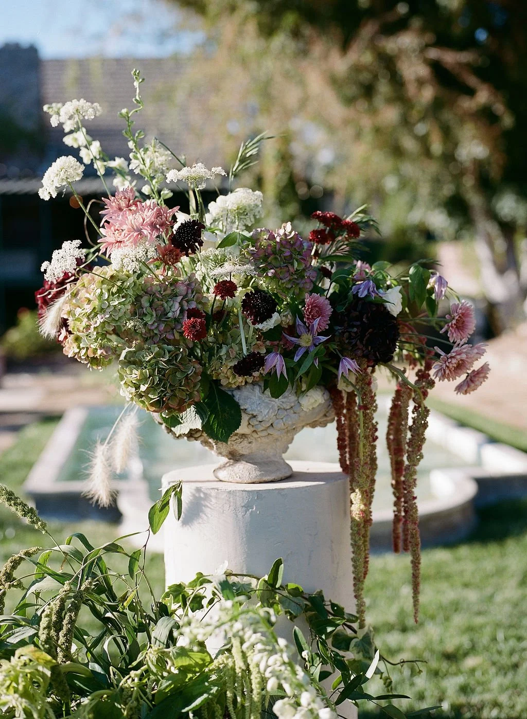 A large floral arrangement of various flowers including hydrangeas, dahlias, and others in a decorative white urn outdoors on a white pedestal, with a blurred background of trees, grass, and a building.