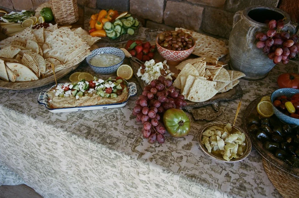 A table set with various foods including pita bread, sliced lemons, grapes, strawberries, assorted vegetables, hummus, feta cheese, olives, and a large ceramic jar of grapes, with a decorative tablecloth and a stone wall in the background.