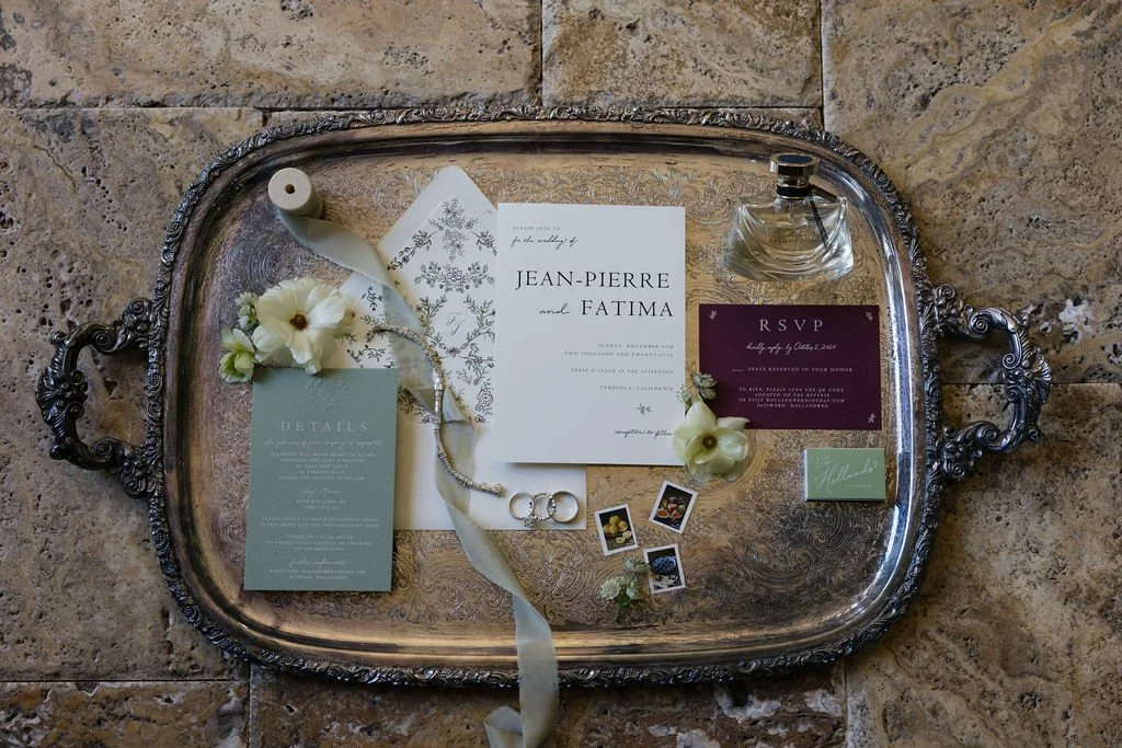 Silver serving tray on a stone floor holds wedding invitations, RSVP card, details card, perfume bottle, flowers, a ribbon, jewelry, Polaroid photos, and a small green card.