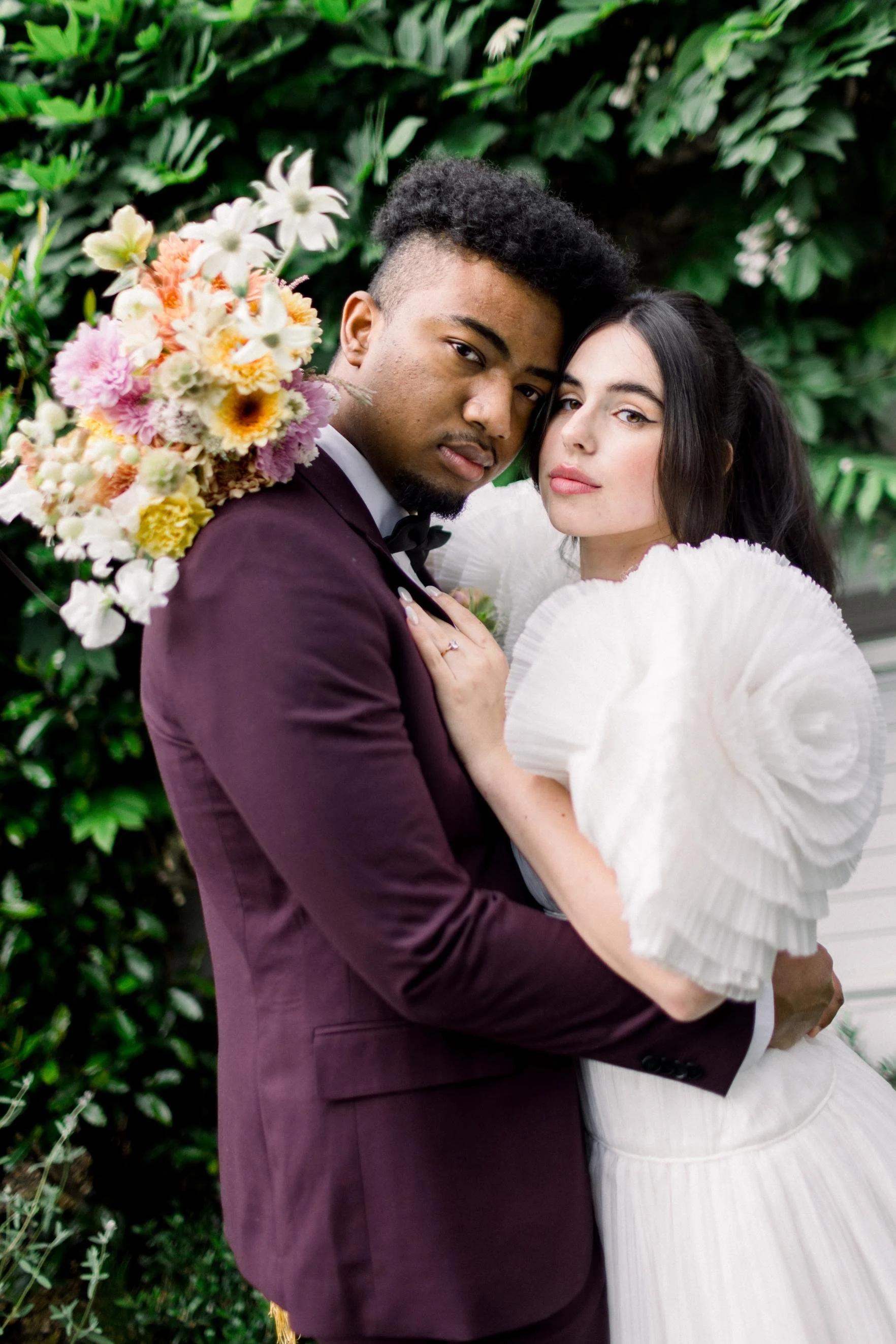 A couple dressed in formal attire posing closely outdoors with lush green foliage in the background. The woman is wearing a white dress with large, puffed sleeves, and the man is in a dark maroon suit and black bow tie. The woman holds a bouquet of c