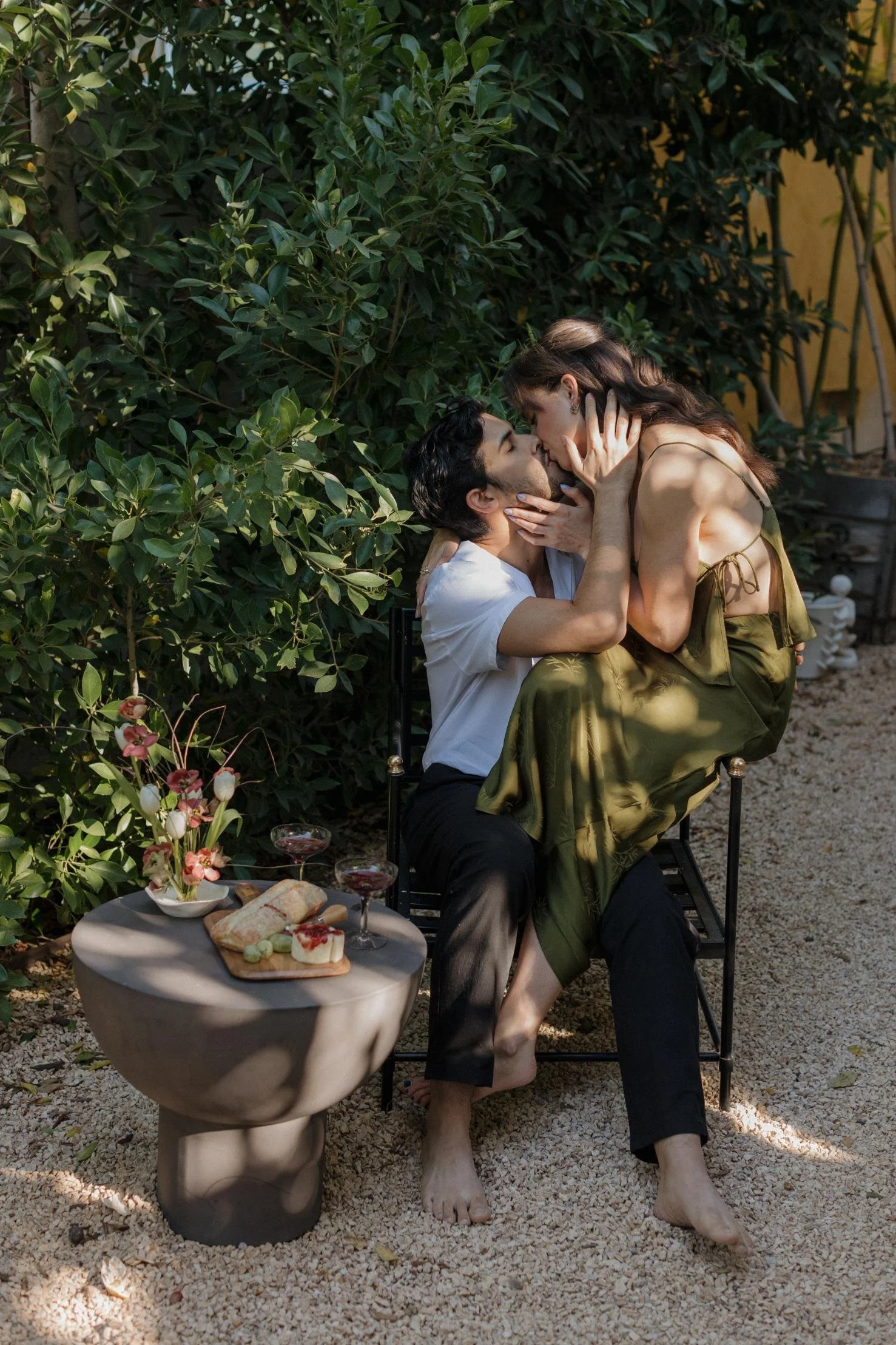 A couple sharing an intimate moment outdoors, sitting on a chair surrounded by lush greenery, with a small table beside them holding flowers, cheese, and drinks.