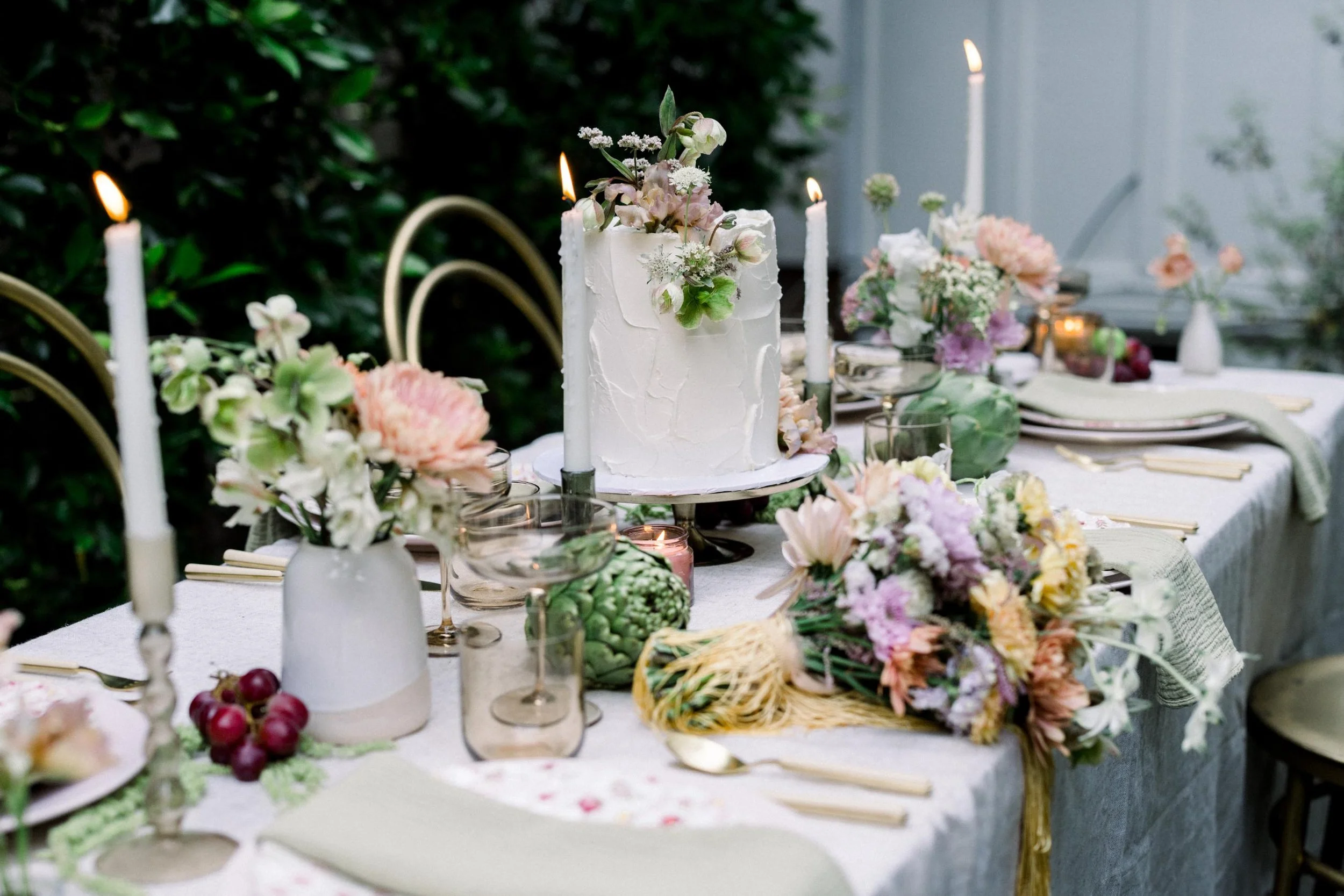 A decorated outdoor dining table with a white cake with floral decorations, surrounded by pink and white flowers, candles, glasses, and plates, set for a celebration.