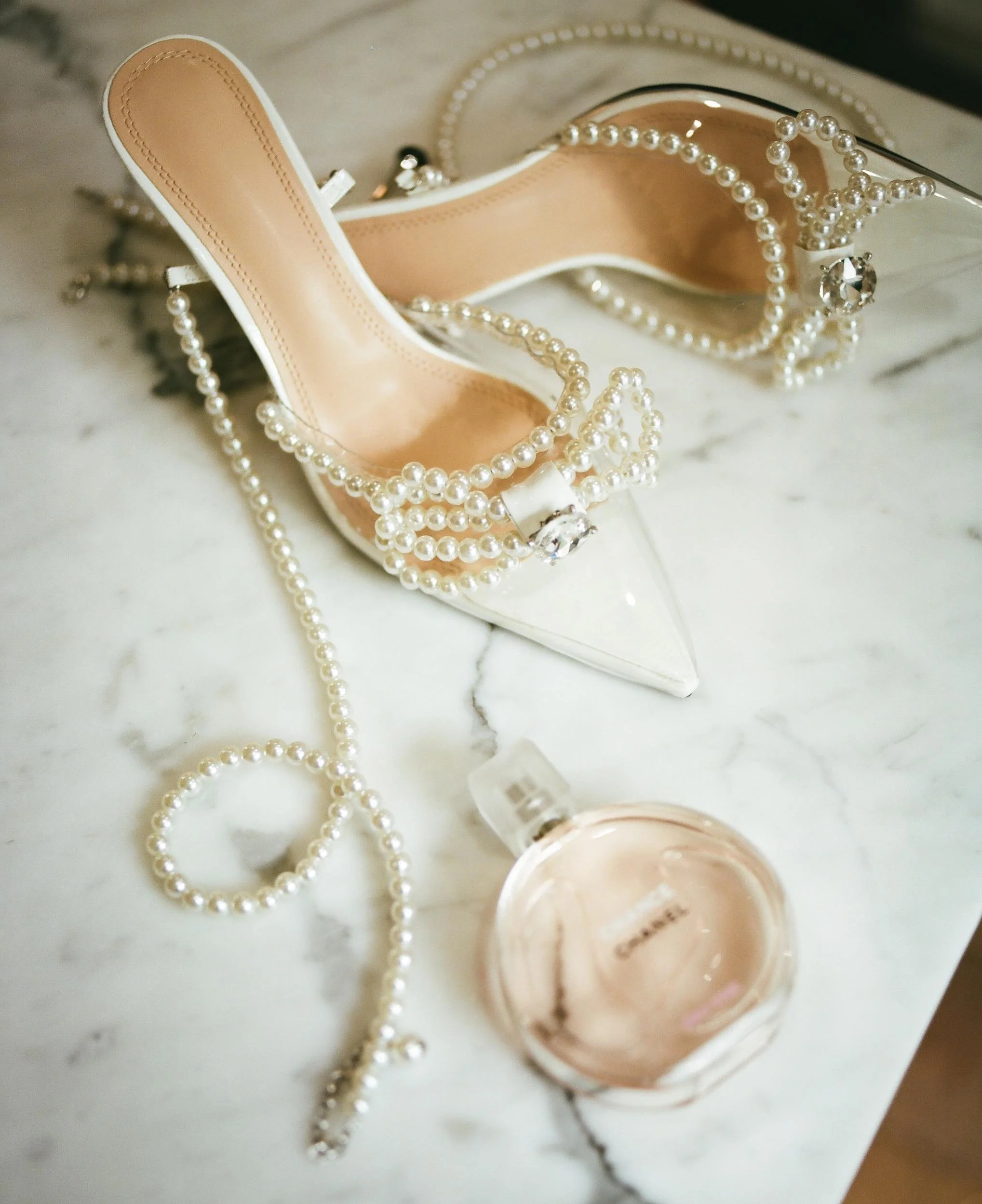 A pair of white high-heeled shoes decorated with pearl strings, a bottle of Chanel perfume, and a pearl bracelet on a white marble surface.