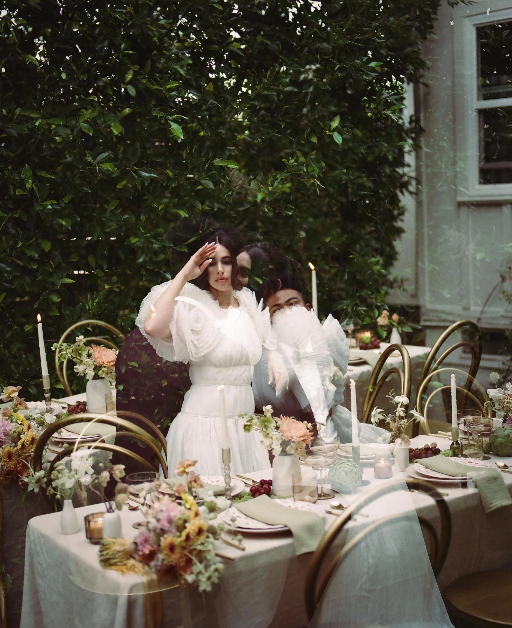 A woman in a white dress standing next to a seated man at an outdoor dinner table decorated with flowers and candles, with lush green foliage in the background.