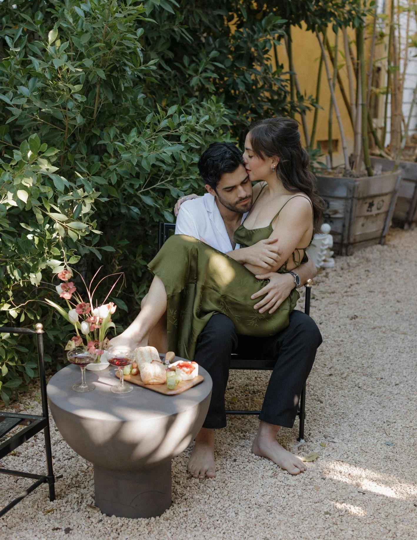 A man and woman sitting closely on a garden bench, with the woman sitting on the man's lap, sharing an intimate moment. The woman is wearing a green dress, and the man is in a white shirt and black pants. In front of them, there is a small round tabl