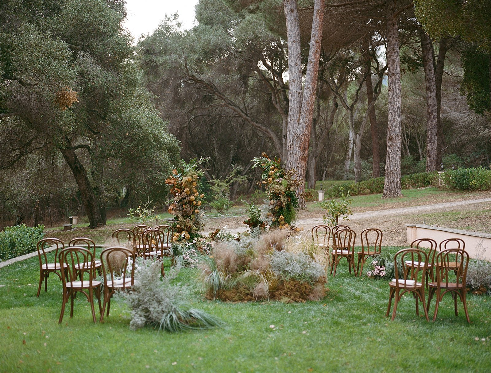 Outdoor wedding ceremony setup with wooden chairs arranged in a semi-circle around a floral and foliage altar, in a lush green garden surrounded by tall trees.