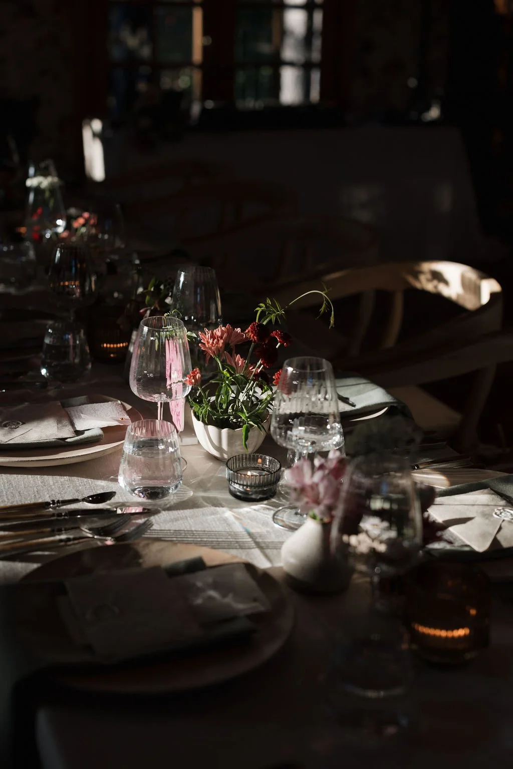 Elegant indoor dining setup with a long banquet table decorated with candles and floral arrangements, situated under an arched stone structure with string lights and greenery.