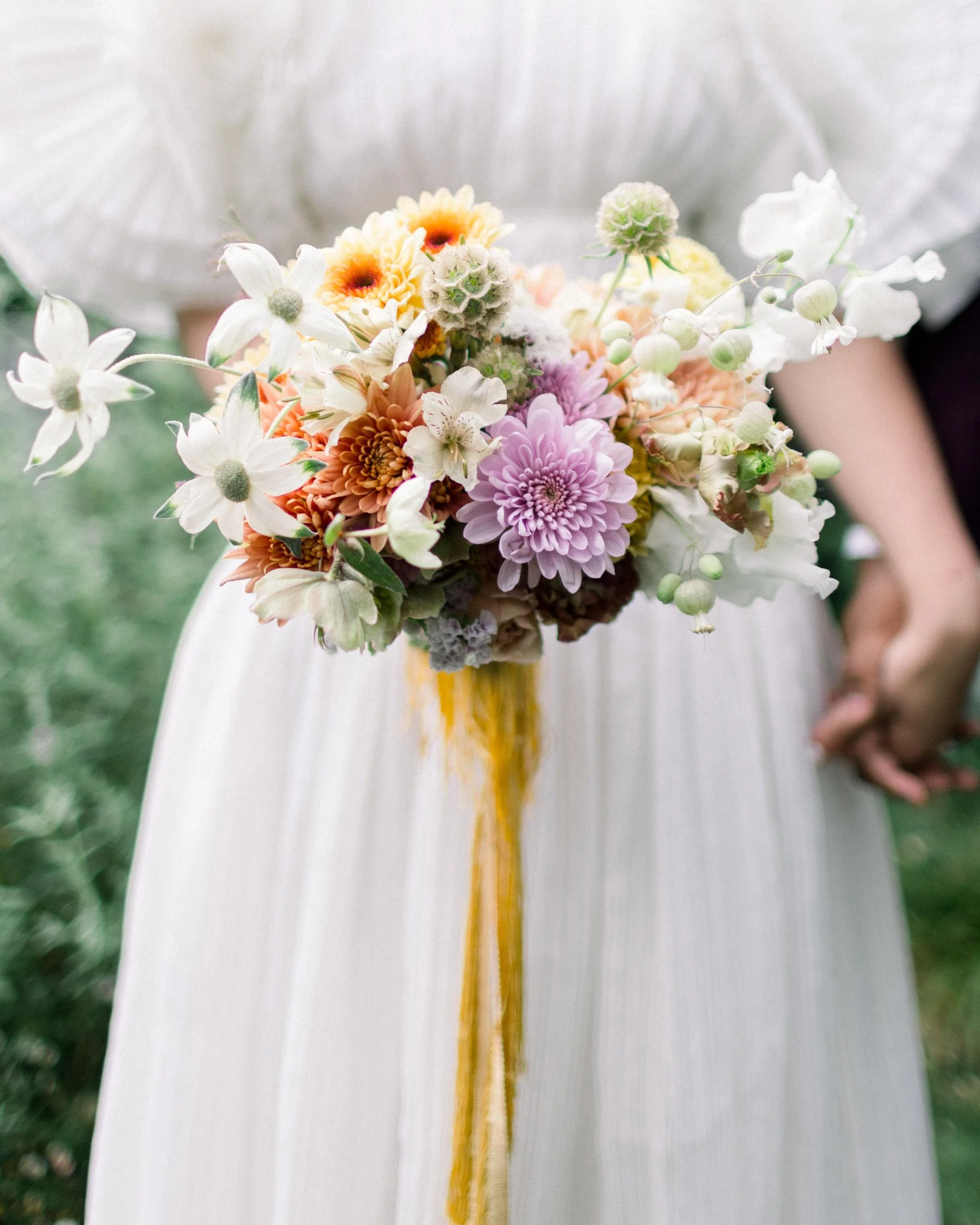 A person in a white dress holding a colorful bouquet of flowers with purple, peach, white, and yellow blooms, some buds, and green foliage.