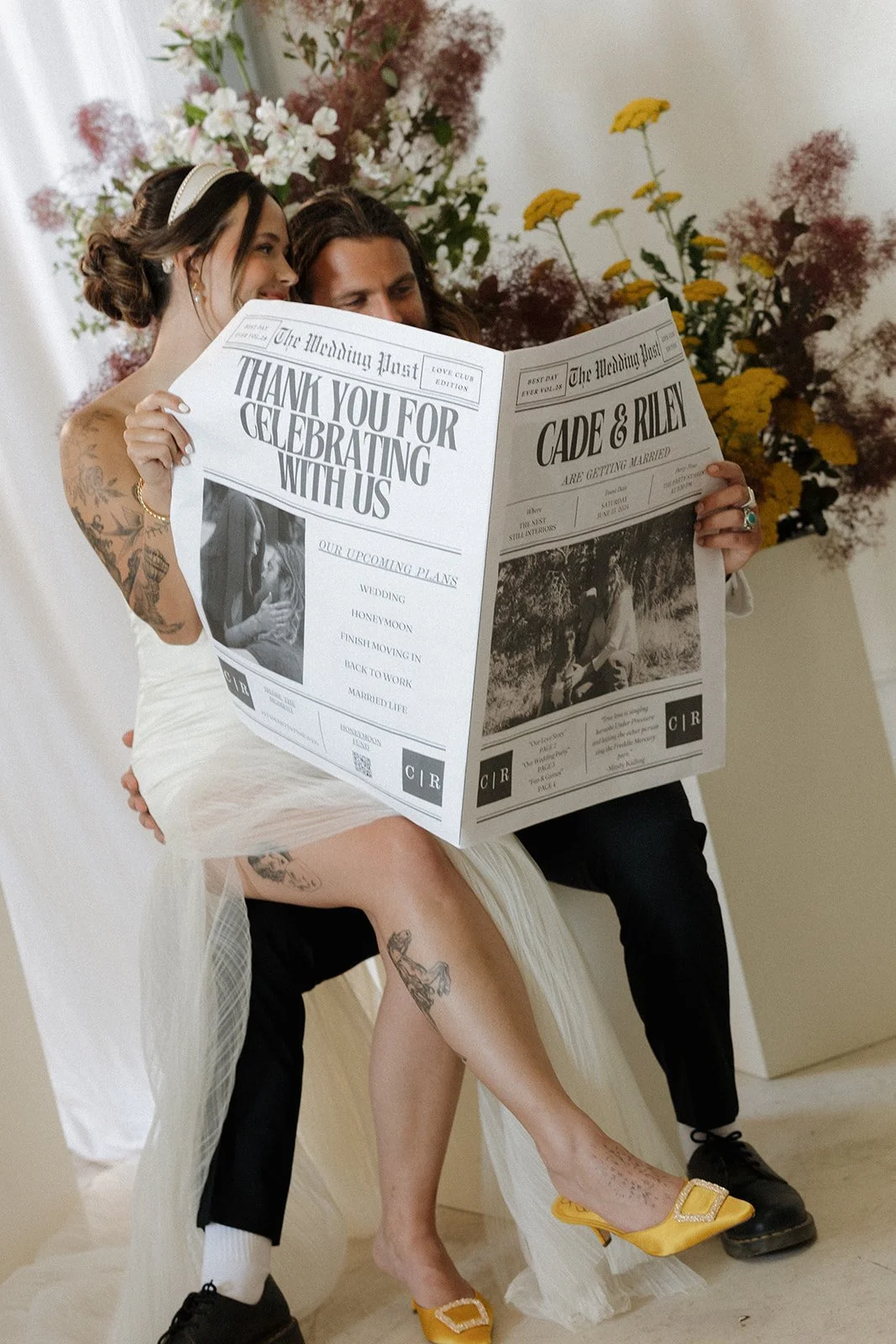 A bride and groom sitting and reading a newspaper at their wedding, with the bride wearing yellow heels and the groom in black shoes, behind a floral arrangement.