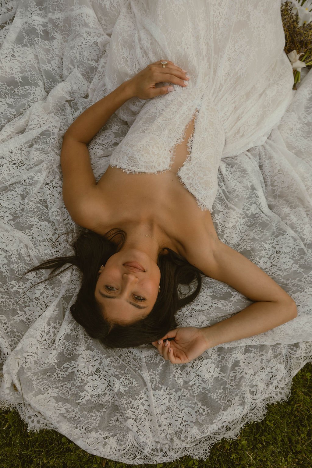 A woman lying on a lace-covered bed outdoors, wearing a white lace dress, smiling, with part of her face turned toward the camera.